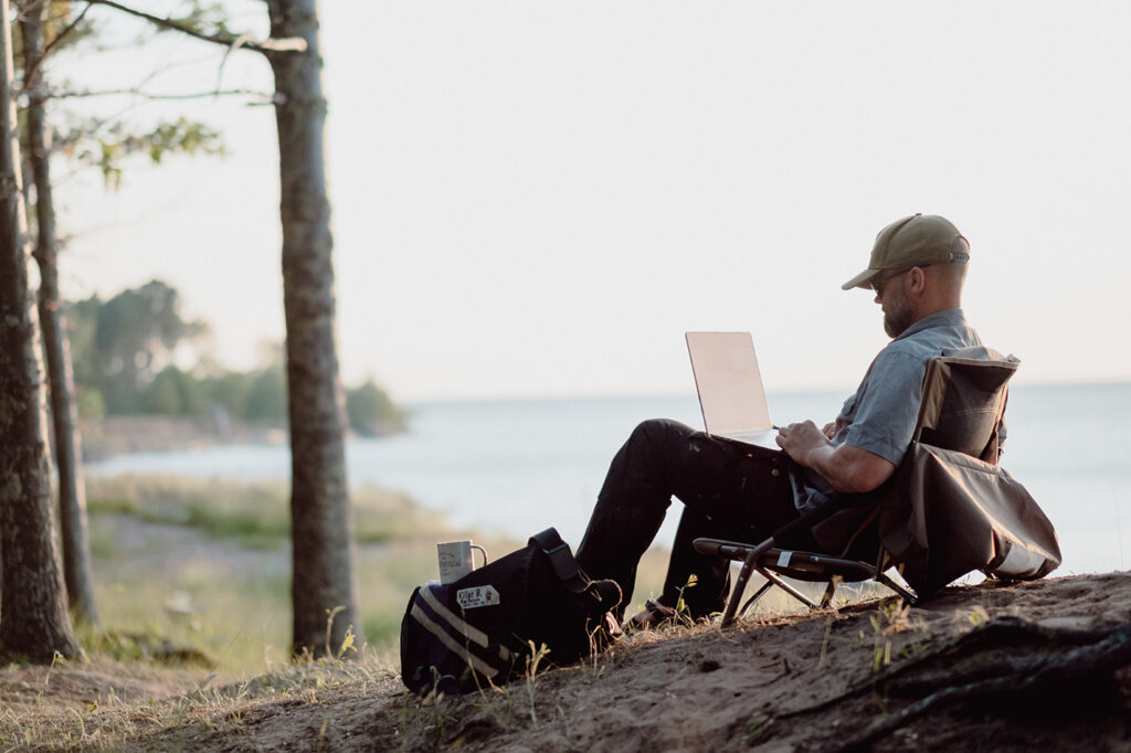 man wearing a ball cap sitting in a camp chair working on a laptop with the lake in the background